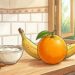 Una naranja brillante y un plátano maduro sobre una encimera de madera en una cocina luminosa, acompañados de un tazón pequeño de yogur y algunas almendras enteras.
