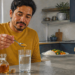 Hombre sentado en una cocina vierte una cucharada de vinagre en un vaso de agua, junto a un plato con arroz y verduras.