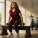 Mujer adulta realiza un ejercicio de fuerza sentada en un banco, con una mancuerna en cada mano, en una sala luminosa con bicicleta estática al fondo.