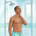 Un hombre joven de piel morena disfrutando de una ducha refrescante en un baño luminoso, con agua cayendo sobre sus hombros, representando la sensación de alivio frente al calor intenso.