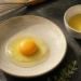 Dos huevos crudos con la clara transparente y la yema intacta servidos en cuencos, junto a un batidor de cocina y hierbas frescas.