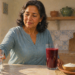 Mujer adulta tomando un vaso de agua en una cocina, junto a una bebida roja y un plato con alimentos.