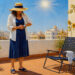 Mujer con sombrero de ala ancha mira su reloj en una terraza soleada, con el sol en lo alto, plantas en macetas, una silla y un vaso de agua.