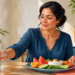 Mujer adulta toma un vaso de agua junto a un plato con sandía, naranja, pepino, tomate, hojas verdes y yogur en una cocina luminosa.
