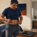 Hombre adulto mide el perímetro de su abdomen con una cinta métrica, junto a una báscula digital y un tensiómetro en el dormitorio.