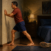Hombre adulto estira la pantorrilla apoyando las manos en una pared del dormitorio durante la noche, con unas zapatillas deportivas junto a la cama.
