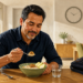 Hombre sentado a la mesa desayuna un cuenco de avena con nueces y manzana, junto a un vaso de agua y un maletín.