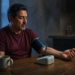 Hombre mide su presión arterial con un tensiómetro automático en una cocina al amanecer, con una taza de café y una copa de vino al fondo.