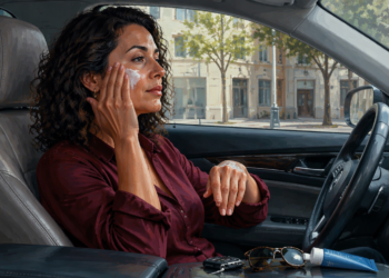 Mujer sentada en el interior de un automóvil aplica crema en el rostro mientras recibe luz solar a través de la ventanilla.