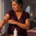 Mujer adulta aplica crema sobre el brazo junto a una ventana iluminada, con una botella cosmética y un cenicero con un cigarrillo sobre la mesa.