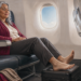 Mujer mayor sentada en un avión con las piernas extendidas y los pies apoyados sobre una maleta, mientras lleva medias de compresión.