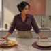 Mujer de pie en una cocina observa dos opciones sobre la encimera: un desayuno preparado y un plato vacío junto a una comida en recipiente.