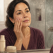 Mujer adulta observa su rostro en el espejo del baño mientras toca la piel de una mejilla, junto a productos de cuidado facial y toallas.