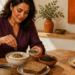 Mujer adulta añade semillas a un cuenco de avena junto a pan integral, legumbres, semillas de chía y una rebanada de pan blanco sobre la mesa.