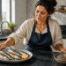 Mujer presenta un plato con pescados enteros asados y limón junto a un cuenco con semillas y nueces sobre la encimera de una cocina.