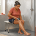 Mujer mayor sentada en un banco dentro de la ducha mientras dirige agua con una ducha de mano sobre una de sus piernas, en un baño adaptado y luminoso.