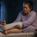 Mujer adulta sentada en la cama durante la noche, usando una toalla para estirar el pie mientras sufre un calambre en los dedos.
