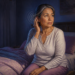 Mujer adulta sentada en la cama durante la noche, tocándose un oído mientras unas ondas circulares representan un zumbido, con auriculares y un ventilador cerca.