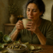 Mujer adulta sostiene una taza humeante junto a varias alcachofas frescas, hojas secas y un cuenco con partes de la planta en una cocina rústica.