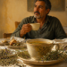 Hombre adulto tomando una infusión en una mesa con una taza grande de hierbas, plantas secas, un frasco de hojas y un pequeño recipiente de vidrio.
