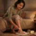 Una mujer de mediana edad sentada en el borde de su cama aplica una crema casera en su talón, con frascos de remedios naturales y una toalla sobre el suelo.