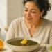 Mujer adulta observando un plato de lentejas acompañado de una rodaja de limón, con una naranja y otras frutas ricas en vitamina C sobre la mesa, en una escena relacionada con la absorción del hierro.