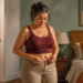 Mujer adulta en un dormitorio intenta abrocharse el pantalón mientras observa la zona abdominal, con una báscula visible al fondo.