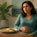 Mujer adulta sentada a la mesa con una mano sobre el abdomen y expresión de malestar, frente a un plato de comida, un vaso de agua y un ordenador portátil.