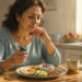 Una mujer madura en su cocina observa pensativa un plato de desayuno saludable mientras sostiene un dispositivo de medición de glucosa en la mano. El plato contiene un huevo frito, rodajas de aguacate, frutos secos y un cuenco con yogur y arándanos.