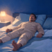Un hombre adulto en pijama acostado boca arriba en una cama dentro de un dormitorio con iluminación nocturna y una lámpara encendida en la mesita de noche.