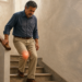 Hombre maduro descendiendo las escaleras con dificultad, apoyándose en la barandilla y sujetándose la rodilla, la cual muestra un efecto de resplandor rojo que indica dolor localizado.