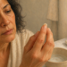 Mujer adulta observando una uña quebradiza mientras la sostiene con los dedos junto a un cuenco con agua y un frasco de ungüento casero, en un baño iluminado, relacionada con el cuidado y fortalecimiento de las uñas.