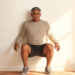Un hombre mayor con ropa deportiva realizando un ejercicio isométrico de pared, manteniendo una sentadilla estática con la espalda apoyada en una pared blanca en una habitación iluminada por el sol.