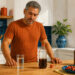 Hombre de mediana edad observa varias bebidas sobre una mesa, entre ellas agua, refresco oscuro y botellas, junto a frutas y un plato con comida.
