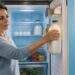 Una mujer madura observa el interior de una nevera abierta mientras sostiene una botella de leche ubicada en el estante de la puerta. En el interior se aprecian huevos, verduras y frascos organizados.