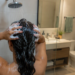 Mujer se lava el cabello con champú bajo la ducha y masajea el cuero cabelludo con abundante espuma.