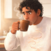 Un hombre joven con cabello rizado y suéter blanco de punto bebe de una taza de café marrón en una cocina soleada, con una expresión de sorpresa y malestar, mientras el vapor sube y una línea de humo ondulada se ve en su pecho, lo que sugiere malestar intestinal. En el fondo, hay una prensa francesa y un frasco de granos de café sobre la encimera.