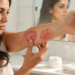 Una mujer examina de cerca dos lesiones cutáneas rojizas distintas en su antebrazo, una de ellas una placa circular escamosa y la otra más difusa e inflamada, en un entorno de baño frente a un espejo, con una expresión de preocupación mientras toca la placa más escamosa.
