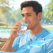 Un hombre con ropa deportiva y sudor en el rostro bebe un vaso de agua sentado en un banco de un parque soleado para hidratarse de forma efectiva.