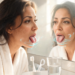 Una mujer en albornoz blanco se mira fijamente en el espejo del baño por la mañana mientras saca la lengua, la cual muestra una capa blanquecina, rodeada de gráficos abstractos de ondas de agua y partículas que sugieren sequedad o bacterias.
