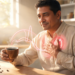 Un hombre sentado en una cocina soleada sostiene una taza de café humeante mientras se lleva la mano al pecho, donde se observa un gráfico luminoso de un latido cardíaco y la silueta de un corazón en tonos rojizos.
