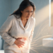 Mujer adulta en un baño sosteniéndose el abdomen inferior con expresión de dolor y malestar pélvico, acompañada de un efecto visual luminoso en la zona del vientre que representa síntomas urinarios como la cistitis.