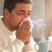 Hombre con suéter blanco limpiándose la nariz con un pañuelo frente a una taza humeante, representando los síntomas de congestión y resfriado.