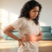 Mujer con expresión de malestar sosteniéndose la espalda baja y el costado en una sala iluminada, mostrando la diferencia entre molestia renal y muscular mediante gráficos luminosos.