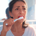 Una mujer con expresión de preocupación se cepilla los dientes frente al espejo, mostrando sangrado en las encías mezclado con la espuma blanca del dentífrico.