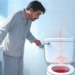 Un hombre en pijama dentro de un baño mira con preocupación el interior de un inodoro con agua de color rojo, representando de forma clara el síntoma de sangre en la orina o hematuria.