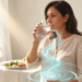 Mujer de perfil en un comedor luminoso bebiendo un vaso de agua antes de comer frente a un plato de comida, ilustrando el hábito de buscar la saciedad.