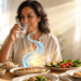 Una mujer de mediana edad sentada a una mesa de comedor bebe un vaso de agua mientras disfruta de una comida saludable que incluye pescado y verduras, con un efecto visual que simboliza la digestión.