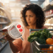 Una mujer en el supermercado analiza la etiqueta de un producto envasado junto a su carrito de compras lleno de frutas y vegetales frescos.