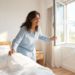 Mujer sonriente en pijama azul abriendo una ventana soleada en su dormitorio para ventilar el ambiente y arreglar su cama con sábanas blancas, ilustrando hábitos para reducir la alergia al polvo en casa.